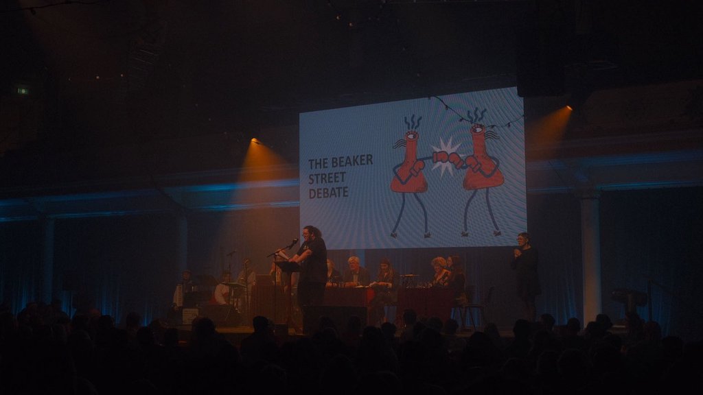 A dark theatre with a packed audience in silhouette. On stage, Tim stands at a lectern with a microphone, a panel of debaters sits behind a long table, and a band with an accordion player waits at the side. A large screen behind them reads The Beaker Street Debate, illustrated with two conical flasks boxing each other? I think that's what it is anyway.