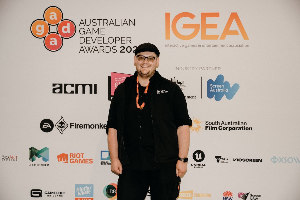 Paris in a black cap and jacket with a Yarn Spinner logo, smiling in front of the Australian Game Developer Awards 2026 sponsor wall.
